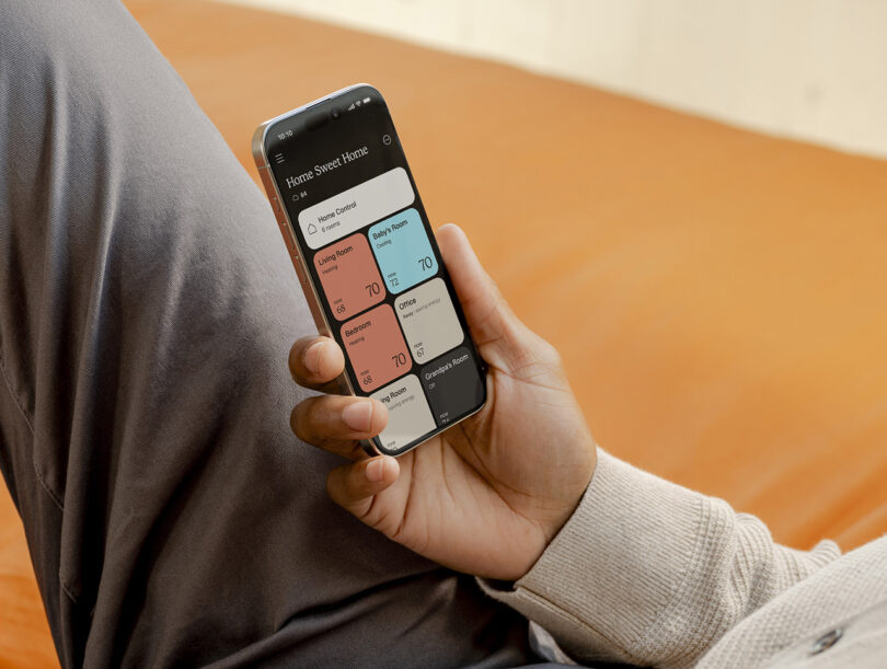Person holding a smartphone displaying a home automation app with controls for various rooms, showing temperature settings of 70 degrees for living room, bedroom, and other areas. The interface seamlessly integrates with your home's climate system to ensure optimal comfort.
