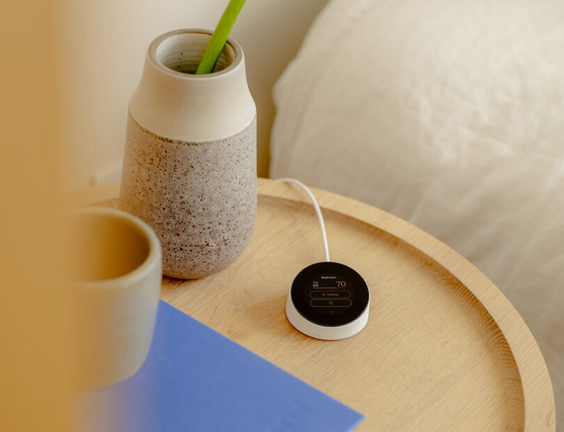 A small round OLED touchscreen remote placed on a wooden table next to a ceramic vase with a green plant and a blue notebook. A white cable is connected to the device connected to control the Quilt heat pump.