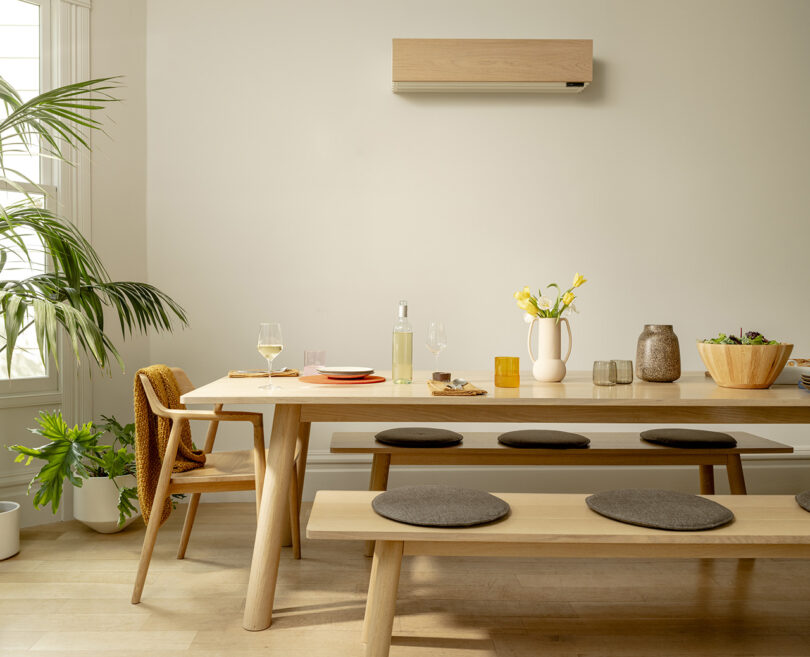 A dining room with a wooden table and benches, a chair, and various decorative items, including plants, a bowl of fruit, and a vase with flowers. For comfort, the room features a modern climate system with a split air conditioner mounted on the wall.