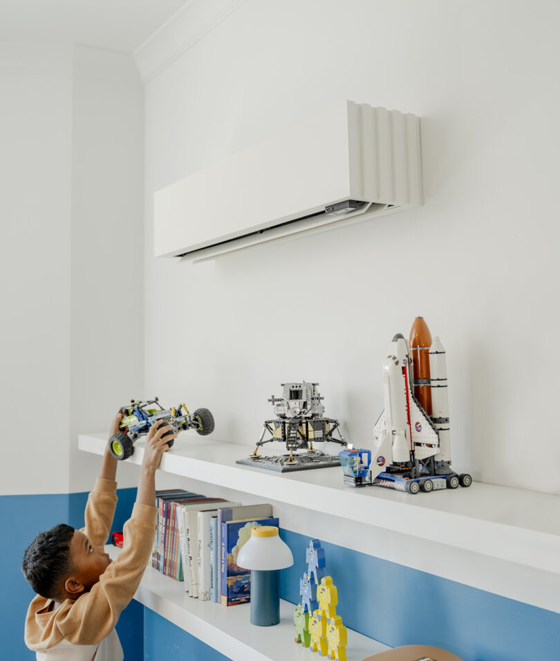 A child holding a toy car stands by a white shelf with books, a lamp, and space-themed models. The shelf is beneath an air conditioning unit from the home's climate system mounted on the wall.