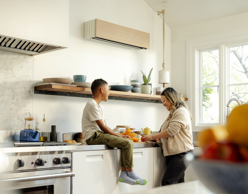 A child sits on a kitchen countertop and a woman stands beside him, both engaged in light conversation. The modern kitchen, equipped with a Quilt heat pump and featuring a window, shelves, and various kitchen items, creates a cozy atmosphere.