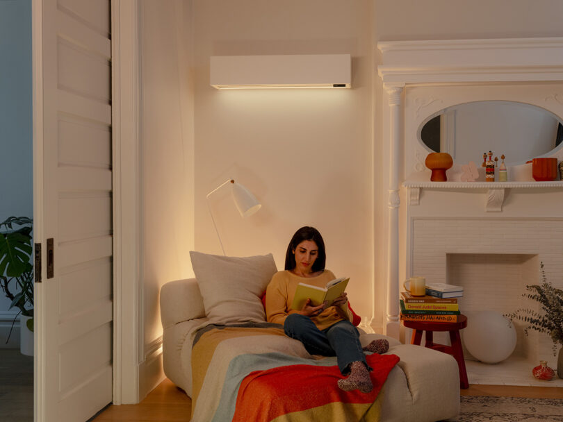 A woman sits on a couch in a cozy, well-lit living room, reading a book. The room features a white fireplace, a side table with books, and a quilt draped over her legs. The climate system maintains the perfect temperature for her comfort.