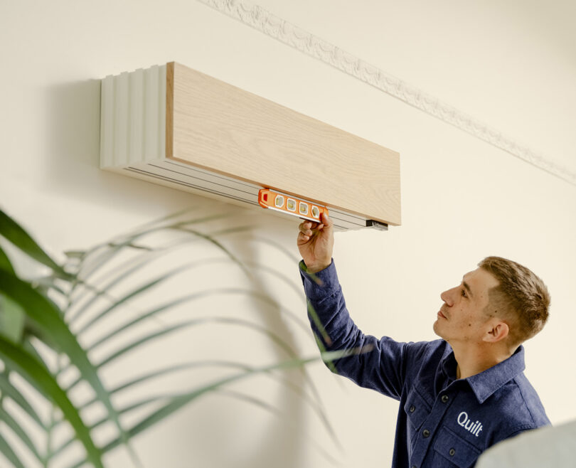 A Quilt technician in a blue shirt using a level tool to check the alignment of a wall-mounted Oak veneer covered mini split unit. A plant is partially visible in the foreground.