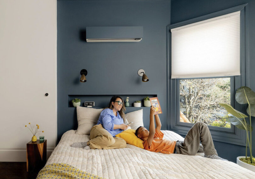 Mom hanging out with son as he reads, each relaxing in bed in bedroom with blue walls and Quilt climate system wall mounted and painted to blend into blue walls.