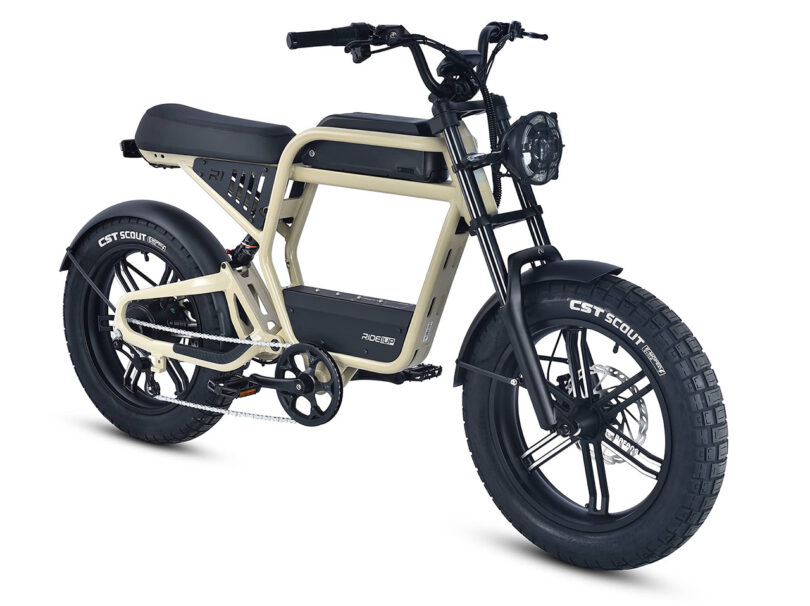The Ride1Up Revv1 eBike showcases a beige frame, large black CST Scout tires, and a rectangular headlight, viewed from a three-quarter front angle. It features a padded seat for extra comfort.