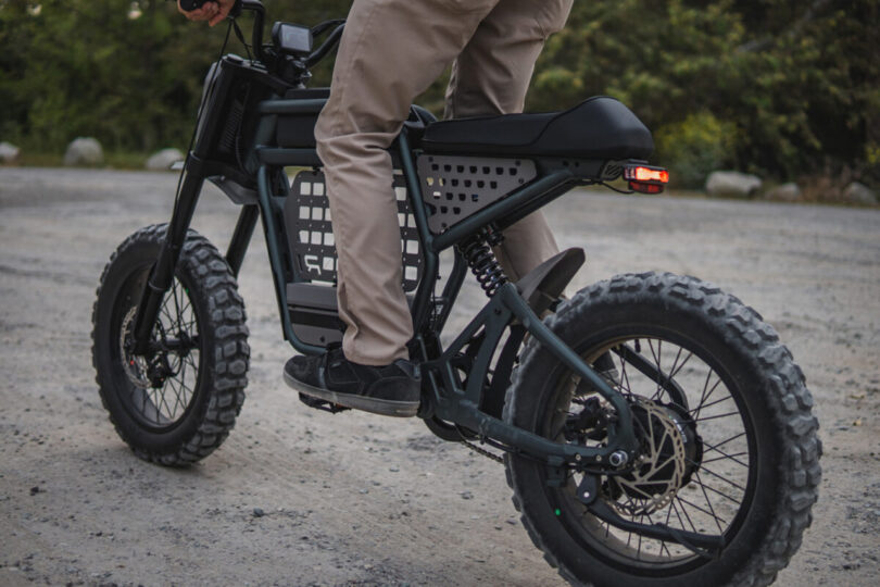 A person riding a black Ride1Up Revv1 DRT eBike on a dirt path, wearing beige pants and dark shoes. The bicycle has thick tires and a rear brake light is illuminated.