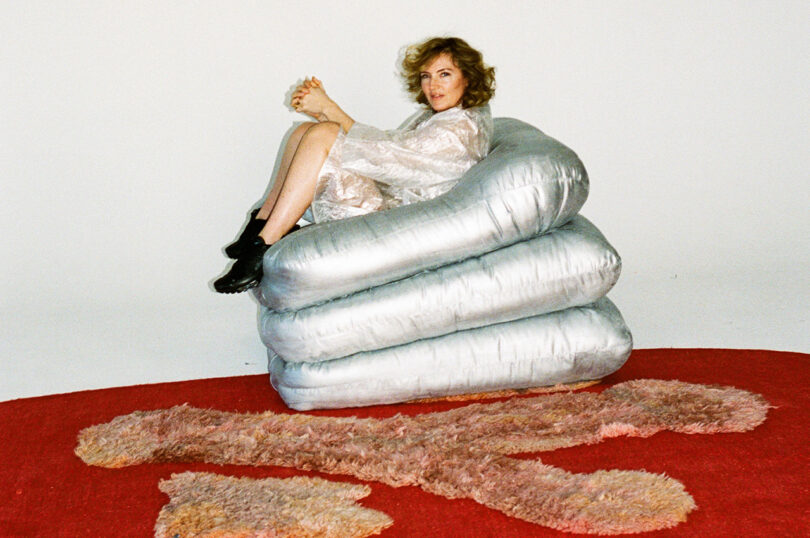 A woman with curly hair, dressed in a silver outfit, sitting atop a stacked, silver structure on a red and pink rug.