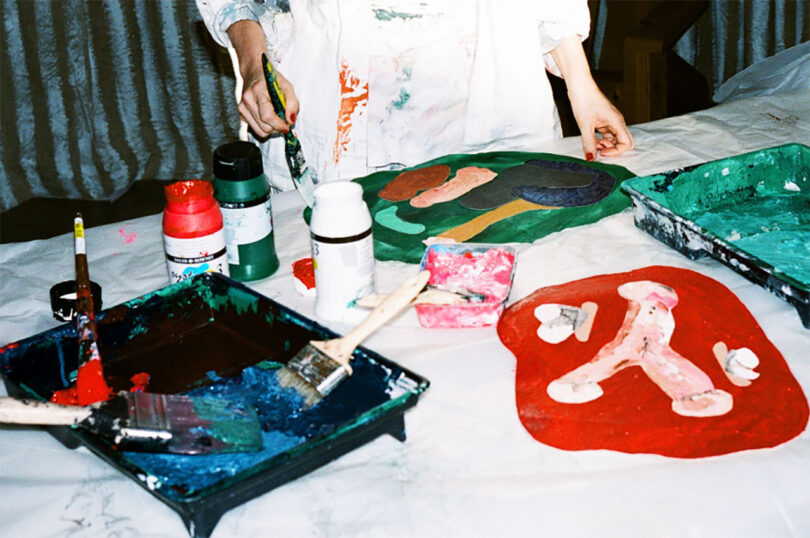 A person working on abstract paintings with a variety of colorful paints and brushes on table.