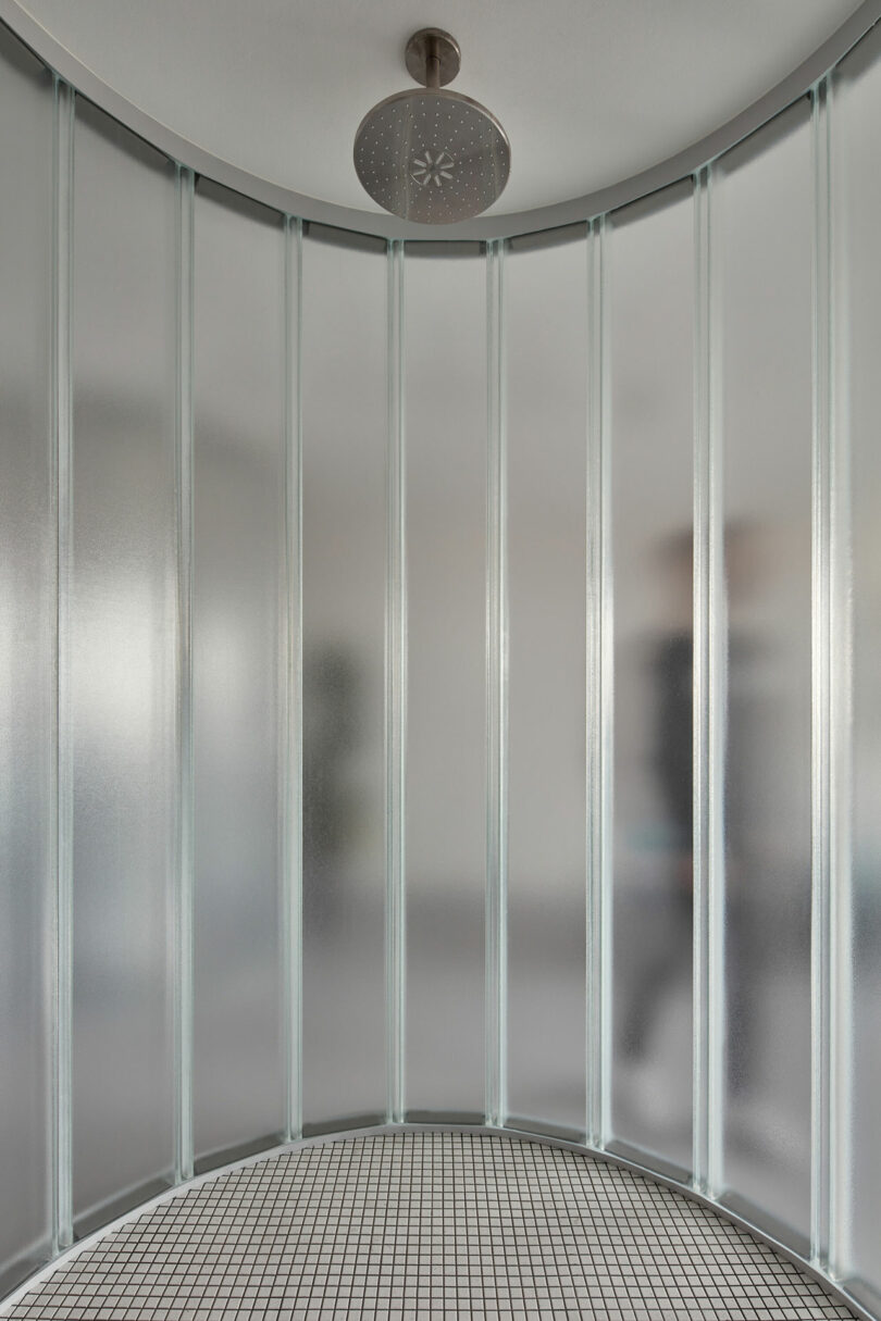 Curved, semi-transparent frosted glass shower enclosure with a large, round overhead showerhead. The floor is tiled with small white and black square tiles.