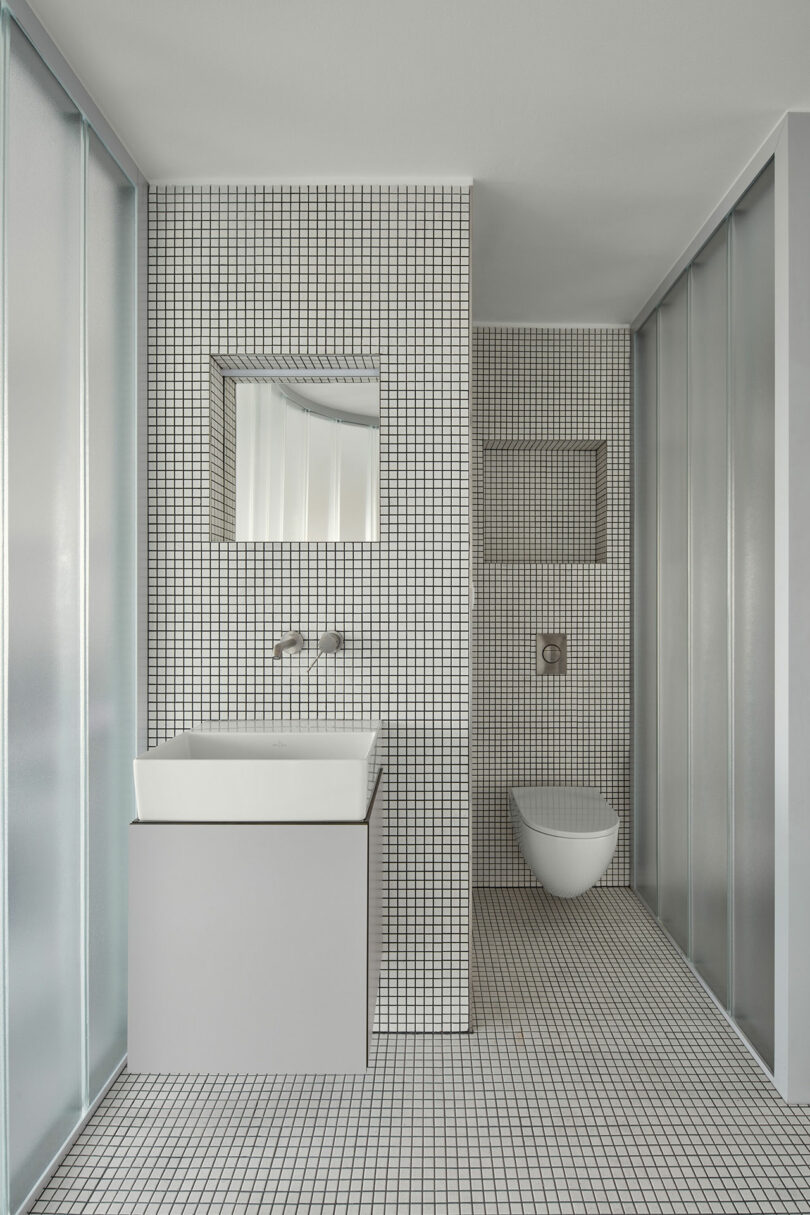 Modern bathroom with white tiled walls and floor, a wall-mounted square sink, recessed mirror, and wall-hung toilet. Frosted glass partitions provide separation within the space.