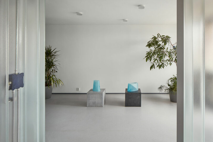 Minimalist room with two concrete blocks, each topped with a blue sculpture. Room is decorated with potted plants in the corners, and has white walls and a light-colored floor.
