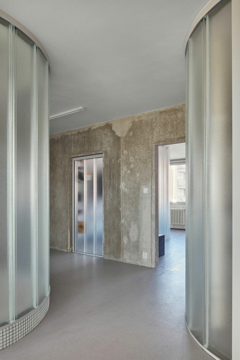 A modern interior featuring frosted glass walls, an elevator with a metallic door, and a partially exposed concrete wall, with large windows in the background letting in natural light.