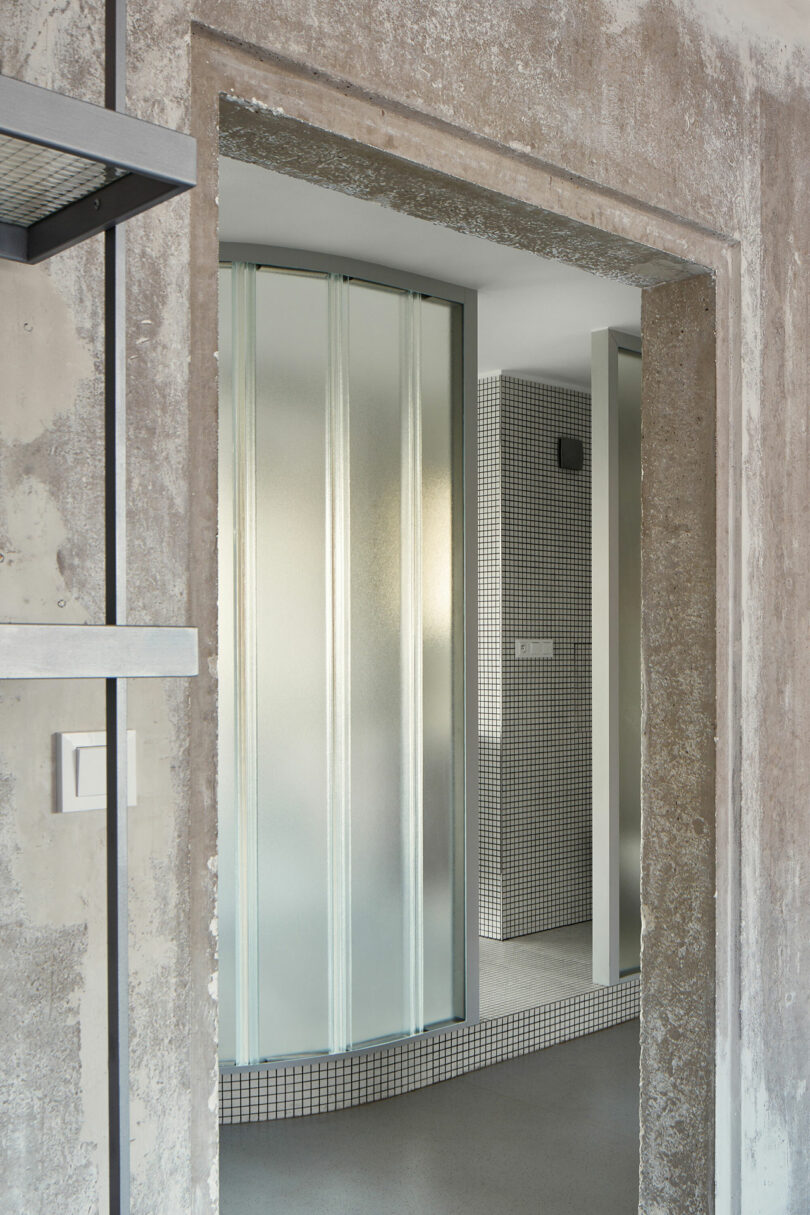 View of a minimalist bathroom with frosted glass partitions, tiled walls, and a rough concrete doorway.