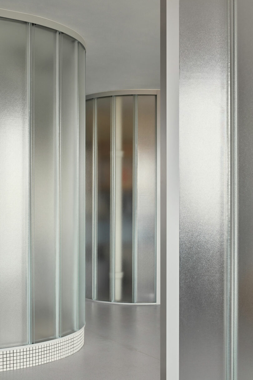A modern interior featuring curved, metallic reflective partition walls and a smooth gray floor. The partitions have a ribbed texture, and the base of the left partition is tiled.
