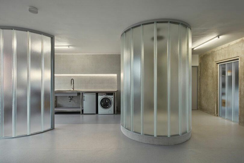 Minimalist modern interior featuring a curved frosted glass wall, a sleek kitchen area with a sink and washing machine, and an elevator on the right.