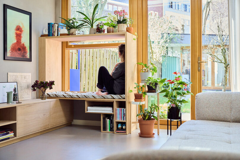 A person sits on a built-in window seat in a modern, plant-filled living room with large windows, surrounded by books and decor.