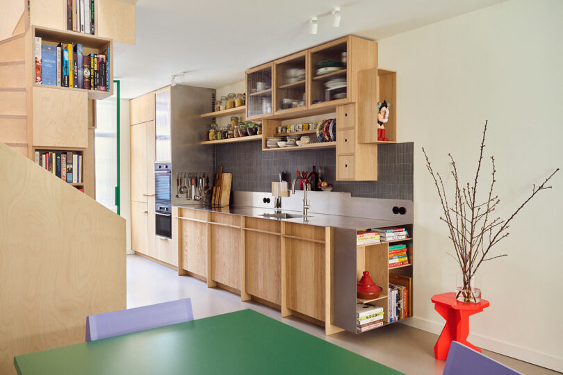 A modern kitchen with wooden cabinets, open shelving, a stainless steel countertop, and a backsplash. Books and decorative items are on the shelves, and a red table with bare branches stands nearby.