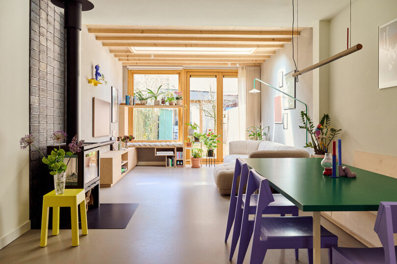 A modern living room with a green dining table, purple chairs, a cozy sofa, and a wood-burning stove. The room is well-lit with natural light from large windows and decorated with plants and contemporary decor.