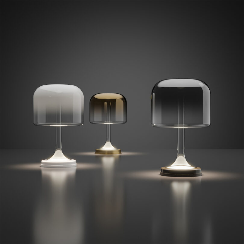 Three modern table lamps with metallic bases and frosted glass shades in white, amber, and smoke colors, displayed on a reflective surface against a dark background