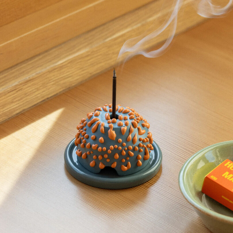 Incense burning in a decorative holder on a wooden table, casting a soft shadow, with sunlight streaming in from the side.
