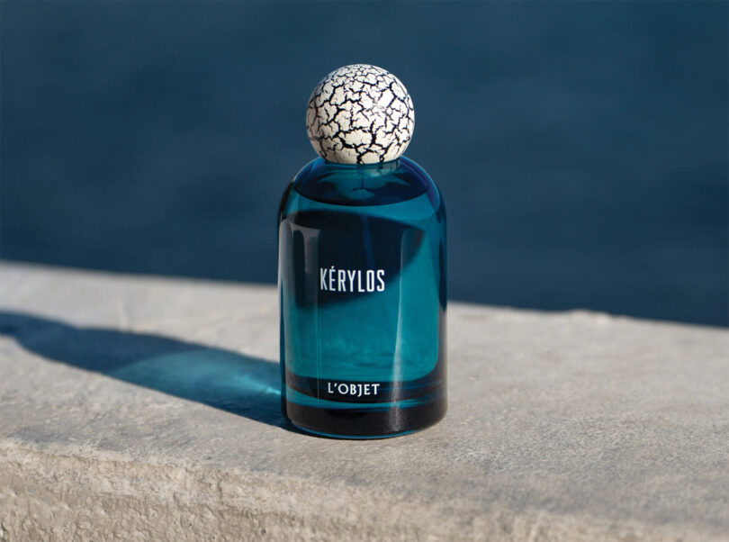 A blue perfume bottle labeled "kerylos l'objet" with a cracked white spherical cap, placed on a stone surface beside water.