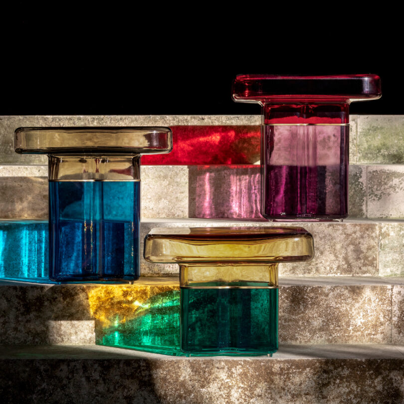 Five transparent, colored murano glass lamps arranged on stone steps, casting vivid shadow patterns in sunlight