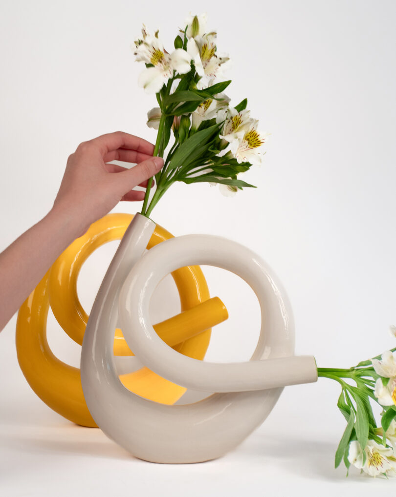 Hand placing white flowers into a modern, curved white vase with a similar yellow vase in the background.
