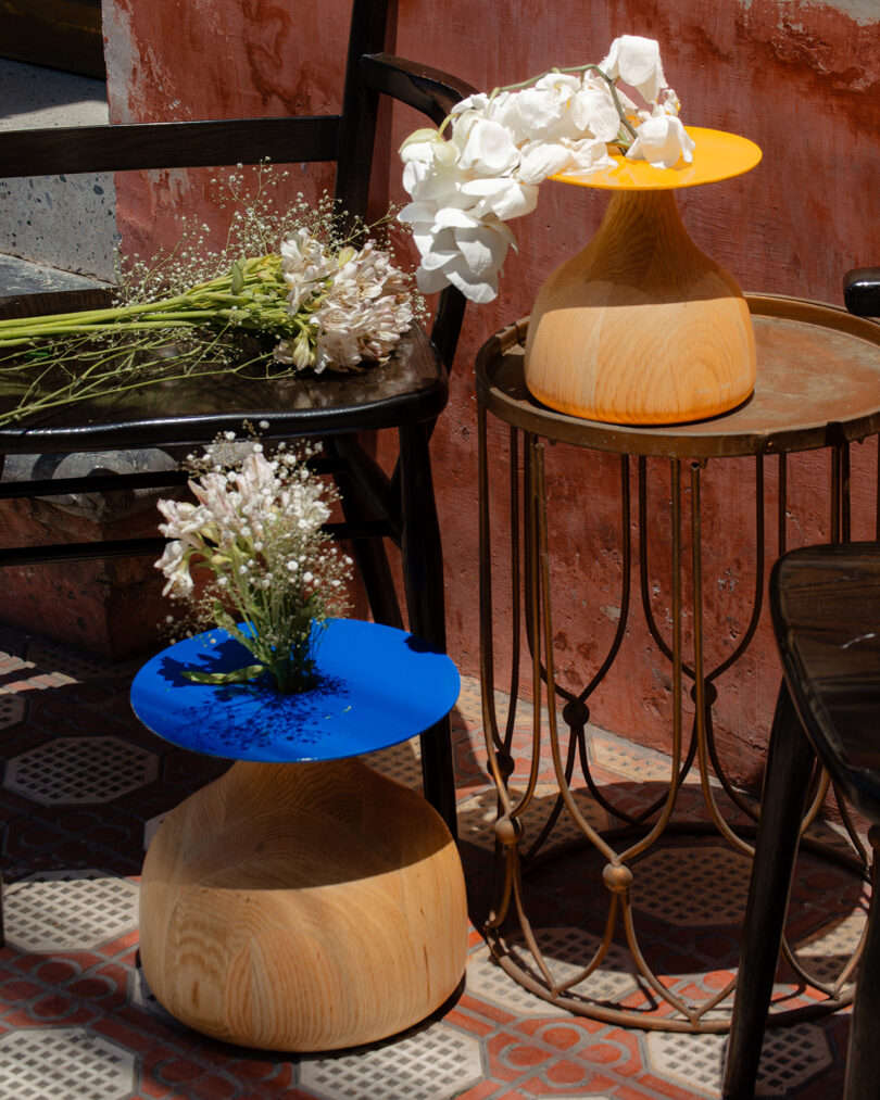 Two wooden stools with bright blue and orange tops hold white flowers and greenery. They are placed near a metal table with a bouquet of white flowers on it, against a rustic wall.
