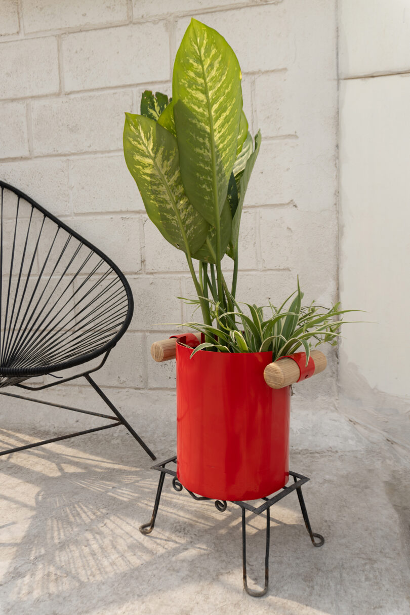A red pot with green plants sits on a black metal stand next to a modern black chair on a concrete surface against a white brick wall.