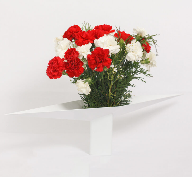 A modern white vase with an angular design holds a bouquet of red and white flowers with leafy green accents.