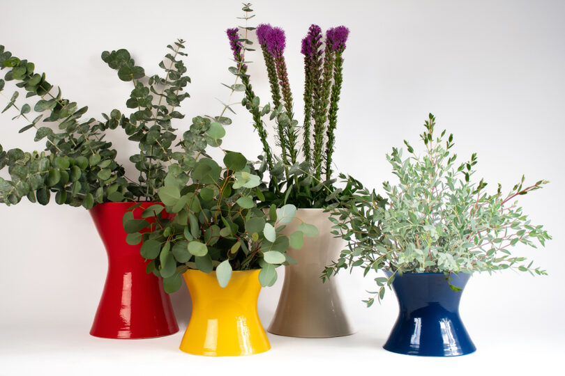 Four colorful hourglass-shaped vases in red, yellow, beige, and blue, each holding different types of plants and flowers, arranged against a plain white background.