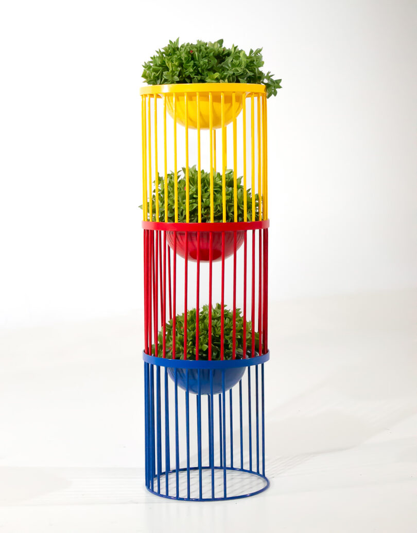 A vertical plant stand with three tiers in yellow, red, and blue, each holding a green potted plant, against a white background.