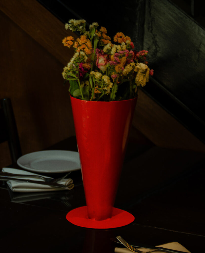 A red conical vase containing a bouquet of colorful flowers is placed on a dark tabletop, with a folded napkin and a white plate in the background.