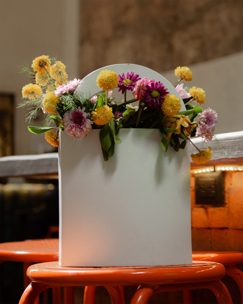 A white vase filled with yellow and pink flowers is placed on an orange stool.