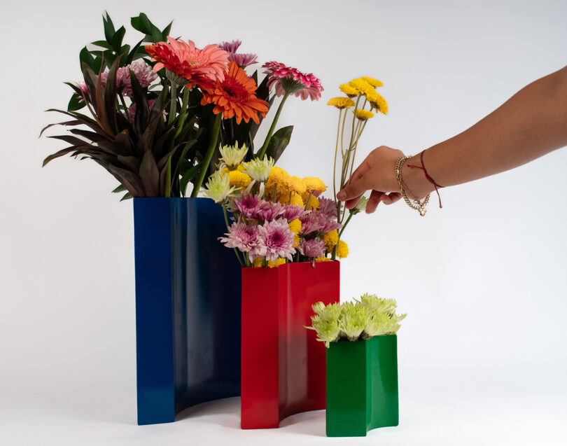 A person's hand arranges flowers in colorful, uniquely designed vases. The vases are blue, red, and green.