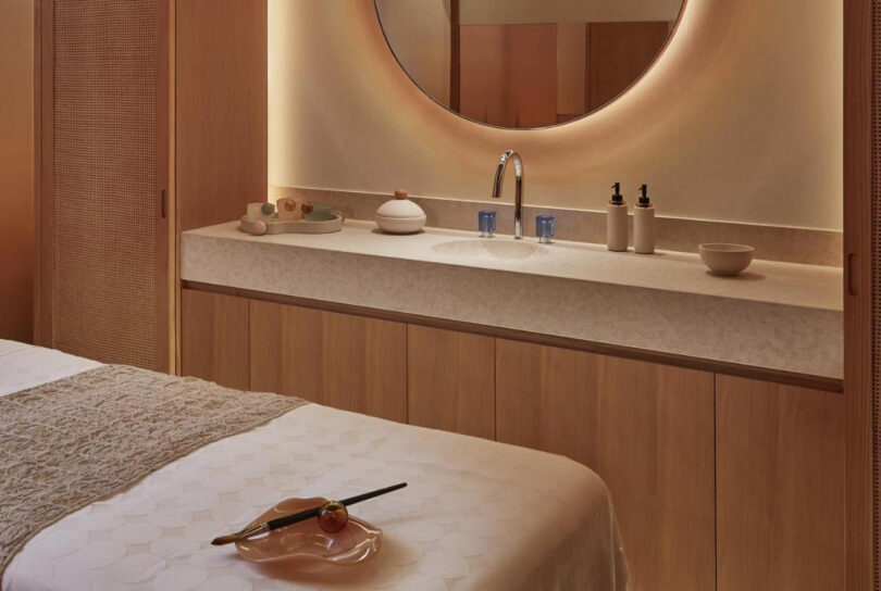A serene spa room at The Emory hotel with a massage bed, a warmly lit round vanity mirror, and wooden cabinetry filled with neatly arranged self-care products.