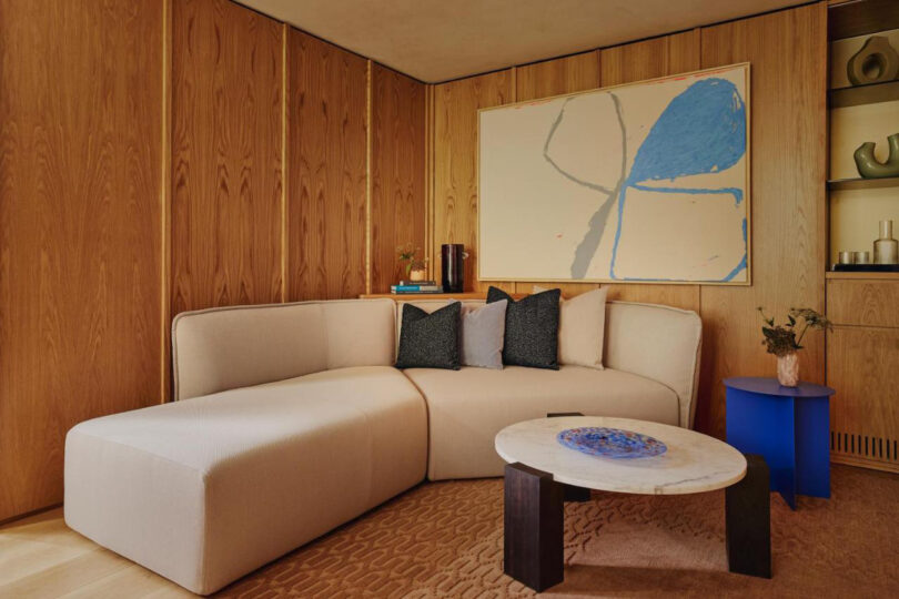 Modern hotel suite with a beige sectional sofa, wooden panel walls, abstract painting, and a contemporary coffee table at The Emory hotel.