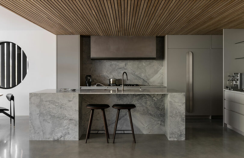Modern kitchen with a marble island, two dark wooden stools, stainless steel appliances, and a slatted wooden ceiling.