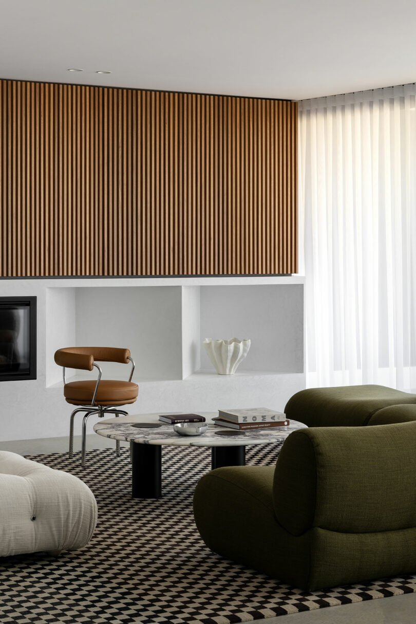 Modern living room with green and white armchairs, a marble coffee table, and a wooden slat accent wall. Books and decor items are on the coffee table and built-in shelves under the wall.