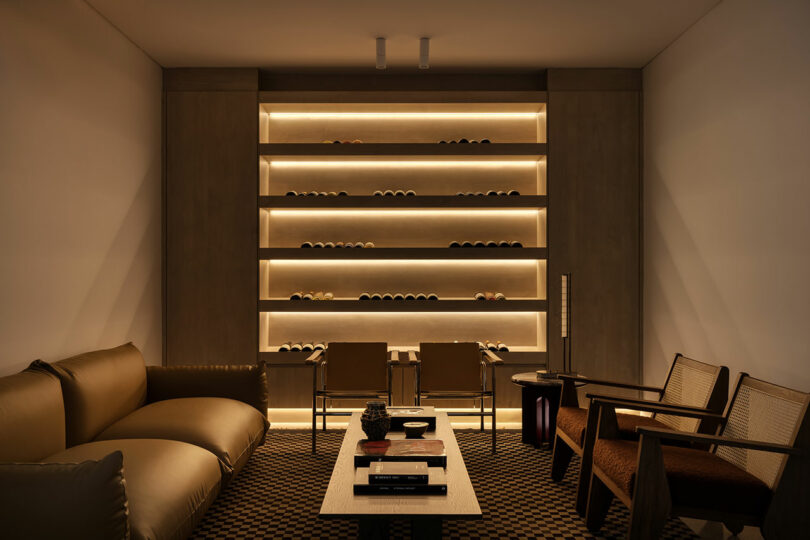 A modern, dimly-lit lounge features a tan sofa, two wooden chairs, a central coffee table with books, and a backlit shelving unit with decor items against the back wall.