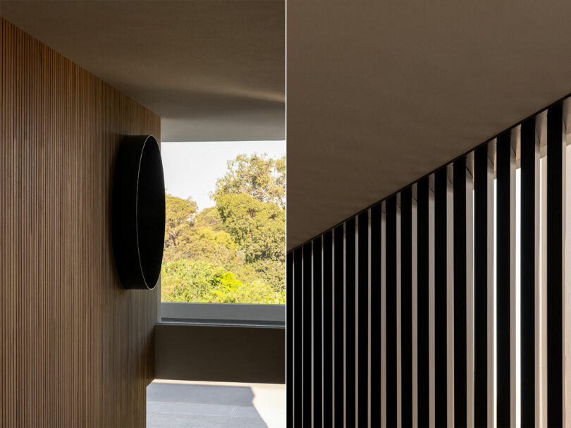 A modern architectural design featuring a circular black wall fixture on a wooden panel and vertical slats creating light and shadow patterns. Trees are visible outside through the window.