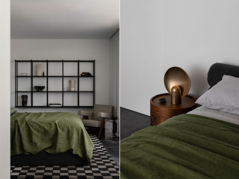 A minimalist bedroom featuring a green bedspread, black metal shelving, and a circular wooden nightstand with a bronze lamp. The shelves hold various decorative items and the room has a neutral color scheme.