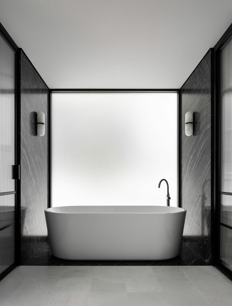 Minimalist bathroom with a freestanding white bathtub, sleek black walls, and two wall-mounted lights on either side. The space features a frosted window behind the tub, providing soft natural light.