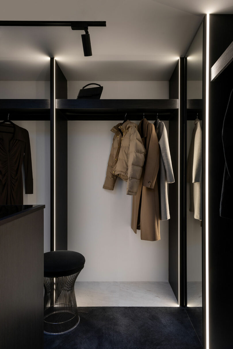 A modern walk-in closet with recessed lighting featuring minimalistic black shelves and hanging spaces. Items include a beige puffer jacket, a tan coat, and a gray garment. A black stool is in the foreground.