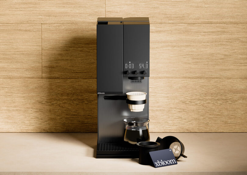 A midnight black xBloom Studio coffee maker with a coffee cup and accessories on a wooden counter against a wooden backdrop.