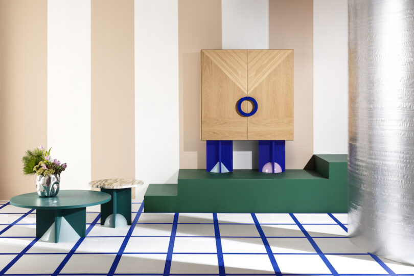 a blue and brown modern cabinet on a green pedestal in a room with striped white and beige walls and square patterned floor