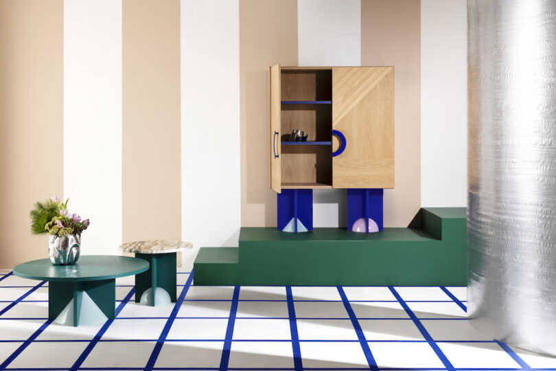 a blue and brown modern cabinet that's opened on a green pedestal in a room with striped white and beige walls and square patterned floor