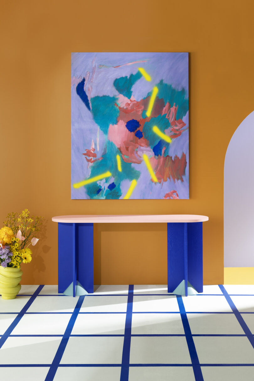 Modern abstract painting on orange wall, blue and white striped floor with a blue console table, and a pot of yellow flowers, arch doorway to the side.