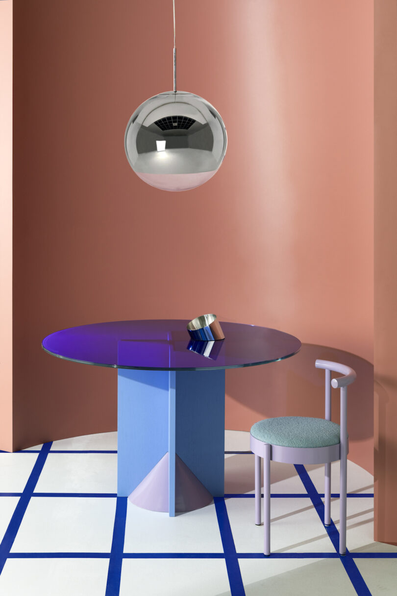 A contemporary interior featuring a blue pedestal table with a purple glass top, a green stool, and a metallic hanging lamp, set against a dual-tone peach wall with blue and white geometric floor design