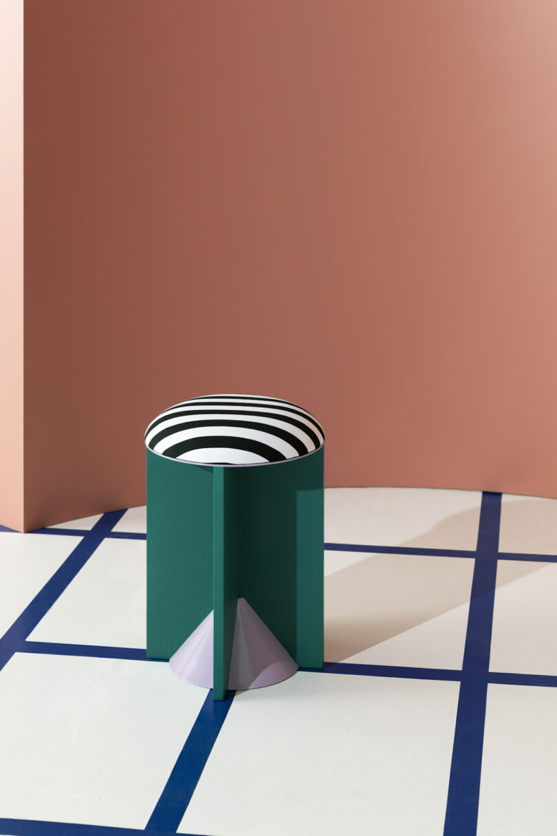A green cylindrical stool with black and white upholstery, positioned on a geometric-patterned floor against a pink wall.